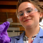 Close up of woman wearing protective eyewear and a lab coat looking at a small square of clear plastic film she's holding.