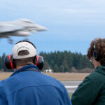 Two men facing away from the camera watch a blurred jet land on an airstrip. The men are both wearing over-ear headphones.