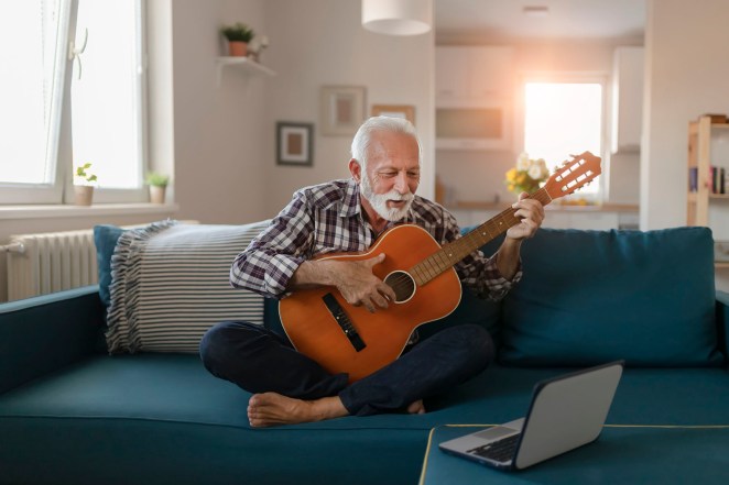 Happy good looking elderly man in a plaid shirt sits on a sofa in the living room and learns to play acoustic guitar online using a Laptop