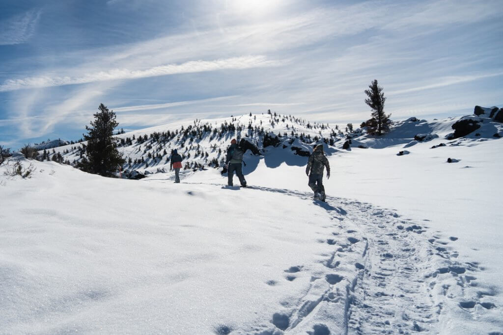 Three people in snow gear, snowshoeing at Craters of the Moon under a sunny sky.