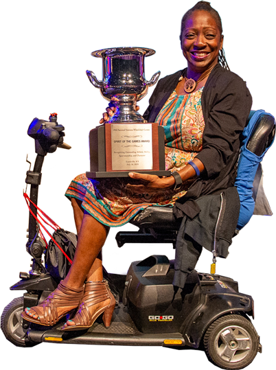 A woman with a beaming smile sits on a mobility scooter while holding a large trophy. She is wearing a colorful patterned dress, a black cardigan, and brown wedge sandals. The trophy inscription reads "Spirit of the Games Award.