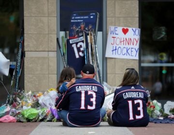 Two people wearing Gaudreau jerseys sit and hold a kid at the memorial