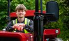 Image shows: Elliot Comerford who has a summer job at Craigtoun Park in St Andrews. Elliot is sitting in the driver's seat of the red tractor called Puffing Billy. He is wearing a high vis vest and black jumper.