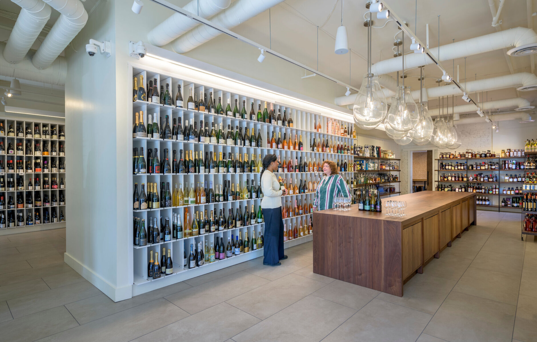 Two people stand underneath dome lights behind a brown wooden table. Both appear to be talking inside the brightly lit wine store