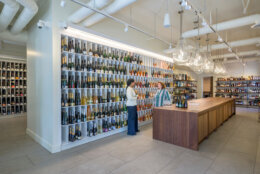 Two people stand underneath dome lights behind a brown wooden table. Both appear to be talking inside the brightly lit wine store