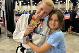 Kelley stands holding a gold medal while wearing her white taekwondo uniform. Her young daughter, wearing a blue shirt, hugs her side.