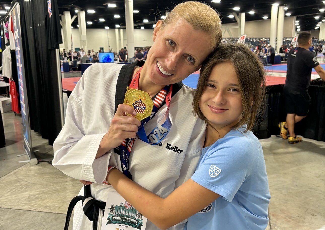 Kelley stands holding a gold medal while wearing her white taekwondo uniform. Her young daughter, wearing a blue shirt, hugs her side.
