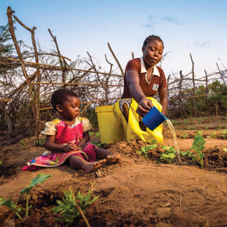 Mother waters her plants in her garden with her toddler sitting next to her.