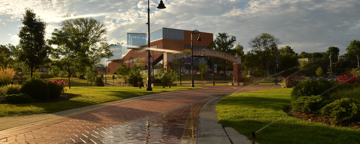 A view of one of Kent State Kent Campus' arches by early morning light. Sprinklers are on and the sun is catching the clouds and the mist and the buildings in the distance.