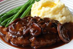 a white plate holding a serving of salisbury steak topped with mushroom gravy, with buttered mashed potatoes and green beans