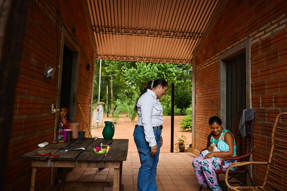 Gladys Nuñez em uma calçada, de frente para uma pessoa que segura um bebê, enquanto uma criança observa de uma porta.