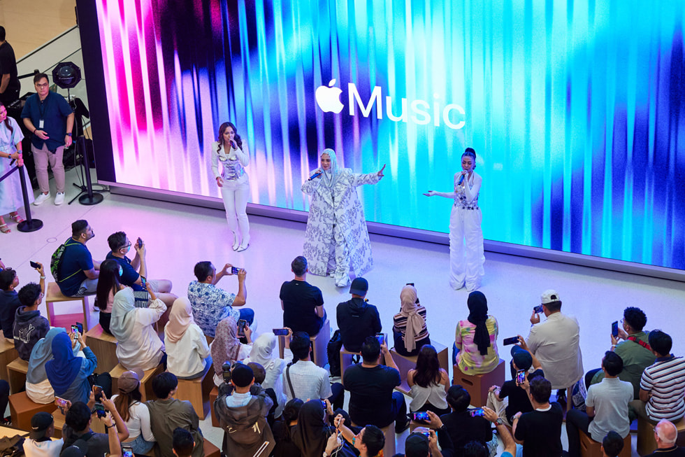 Uma imagem afastada do grupo De Fam se apresentando para os clientes, com o logotipo do Apple Music no telão da parede.