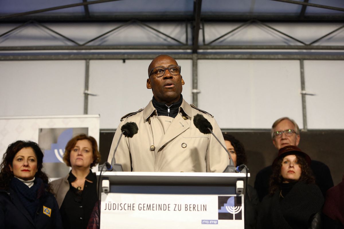 A Black man who wears a trench choat speaking a podium that has on it a menorah and the phrase 'JÜDISCHE GEMEINDE ZU BERLIN.' Several men and women stand around him, looking solemn.