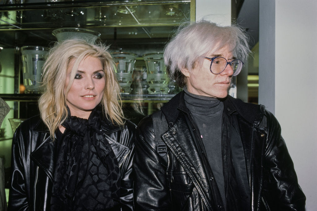 American singer, songwriter, and actress Debbie Harry, wearing a black leather jacket and a black scarf, and American artist Andy Warhol, who wears a black leather jacket over a black turtleneck sweater, attend an AIDS Benefit for St Vincent's Hospital, at Barney's in New York City, New York, 10th November 1986. (Photo by Vinnie Zuffante/Getty Images)