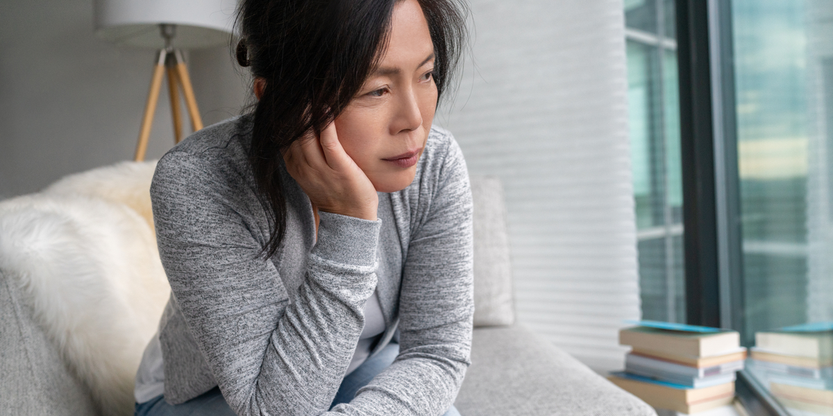 A dispirited-looking woman holds her chin in her hand as she sits on a couch and looks off into the middle distance