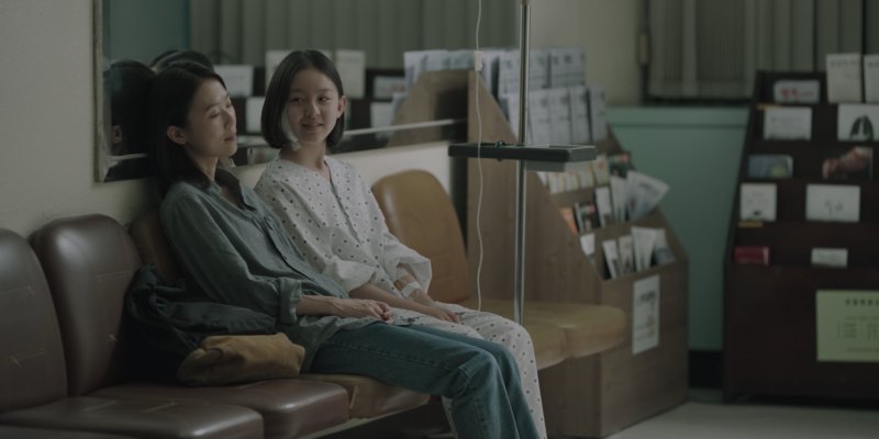 A still from House of Hummingbird. A woman sits next to a girl in a hospital gown and a bandage on the side of her head.