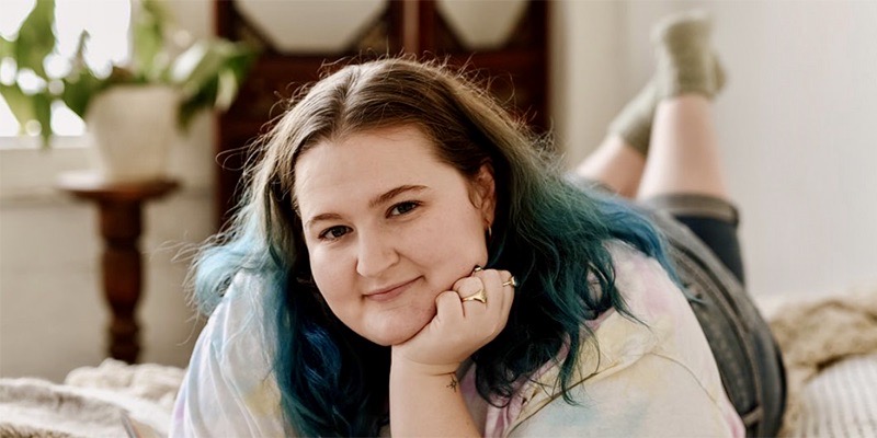 Sophie Carter-Kahn, the host of the podcast "She's All Fat" lays down on her bed with her feet crossed in the air behind her. She has dyed blue tips on her shoulder length brown hair and her face is resting gently on her hand.