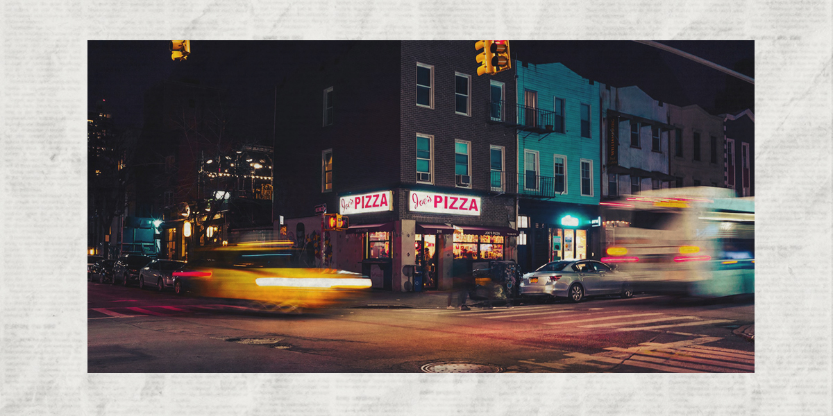 A photo of an NYC intersection at night, with a pizza shop on the corner and traffic blurred running through the intersection under yellow streetlights