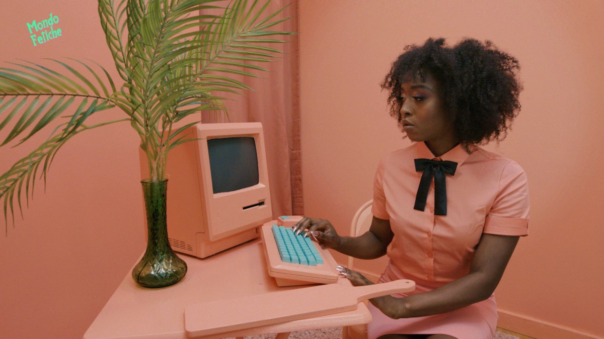 Long nails and lesbian sex: Image shows a Black woman with long nails sitting at a computer in a all peach colored room, typing at the keyboard.