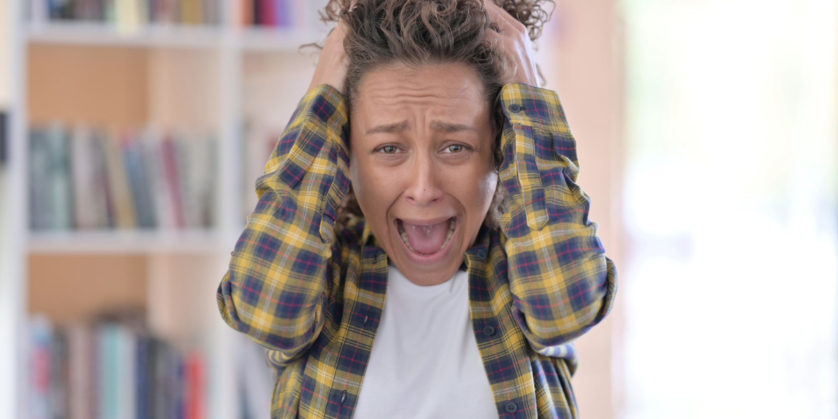 A person with mid-length, curly brown hair wearing a white T-shirt and a blue, red and yellow flannel puts their hands against the sides of their head. Their mouth is open in a frustrated expression.