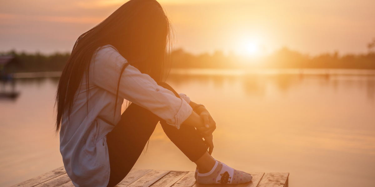 a lonely woman sits with her head between her knees by a beautiful lake