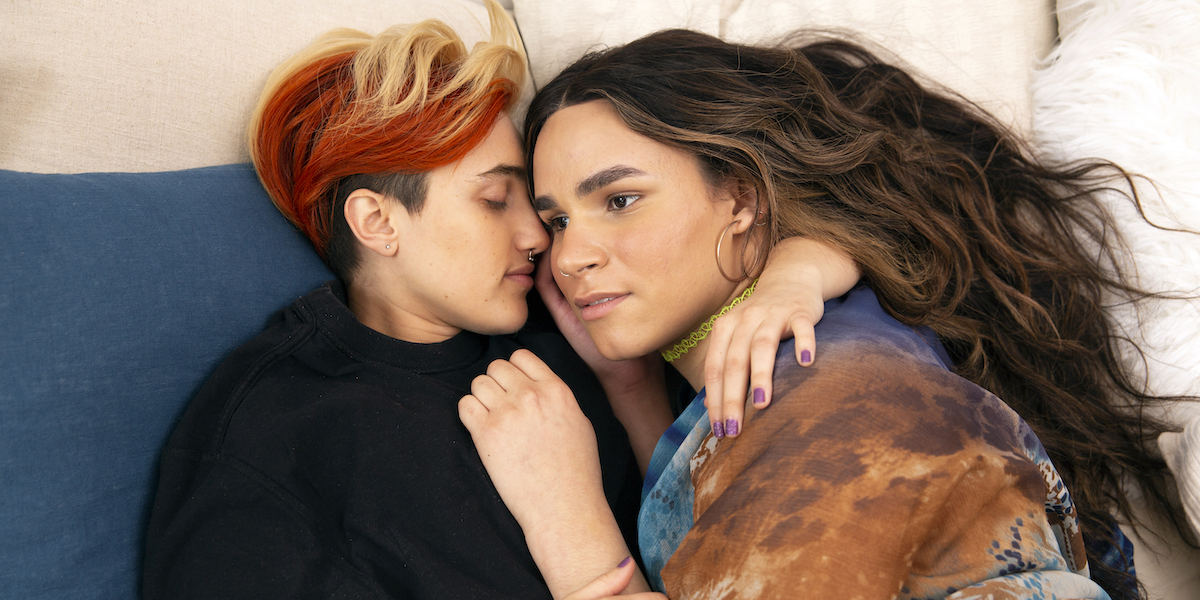 A transmasculine gender-nonconforming person with short blond and bright red hair and a transfeminine non-binary person with long brown wavy hair, waking up together in bed. The person with brown hair is looking into the distance as though thinking about something.