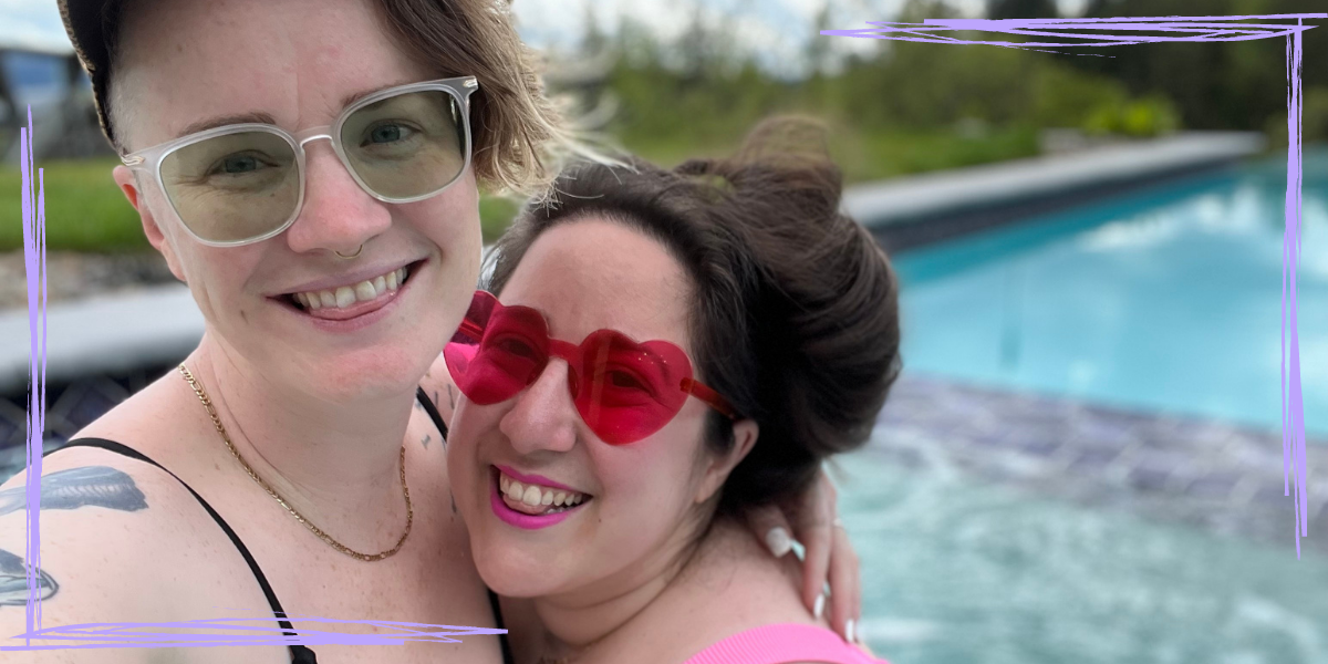 Vanessa and PB pose in front of a pool in bathing suits, wearing sunglasses. Vanessa's sunglasses are red and heart-shaped. PB's are clear with greenish lenses. PB is white and has blonde hair that is long on top and under a hat with a swoop of bangs visible and with shaved sides. Vanessa is also white with long brown hair she is wearing up.