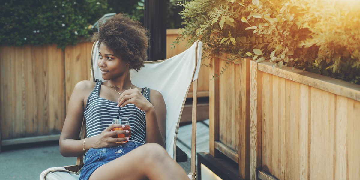 young Black person sitting at an outdoor bar holding a beverage and looking flirtatious