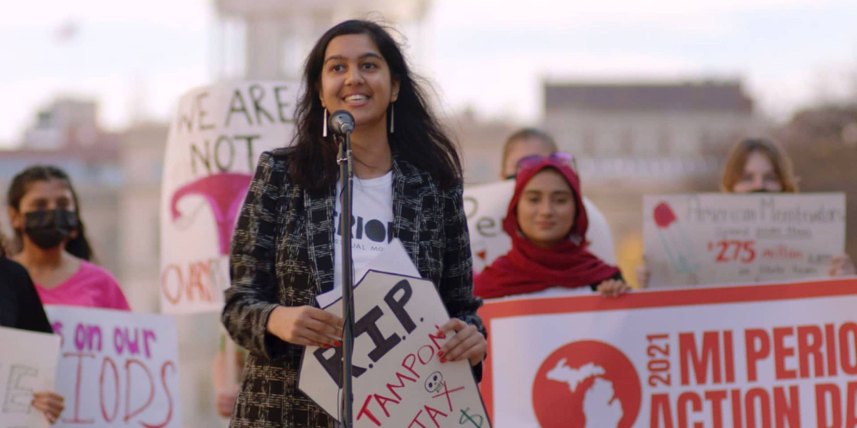 Periodical documentary: a group of young people hold up signs, one says RIP Tampon Tax, and the person holding it speaks at a microphone