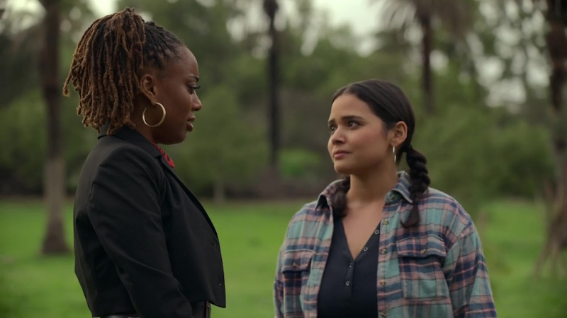 Tracy confronts Malika for half-assing her participation in the retreat activities. Malika is on the left, wearing a black blazer over her flowy red dress. Tracy is on the right, wearing a flannel shirt over a v-neck top.