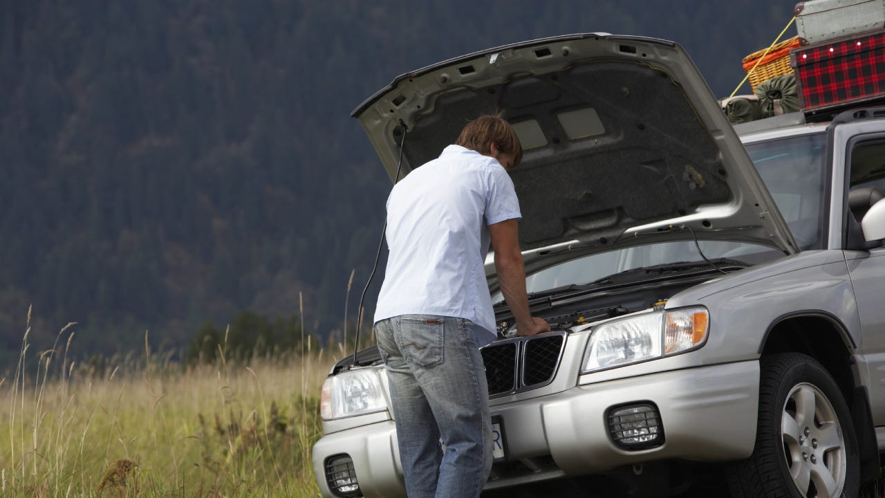 A man stands in front of his car with the hood up trying to get the vehicle fixed.