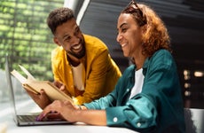 Two university students smile and talk in a library as one holds an open book and the other uses a laptop