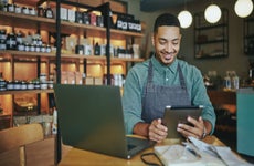 A shop owner wearing an apron smiles as he works on a tablet and laptop in his shop.