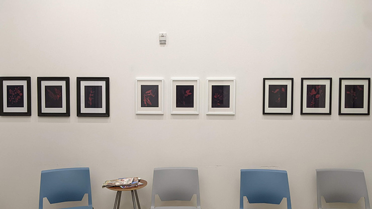 A white wall with 9 framed art pieces above blue chairs.