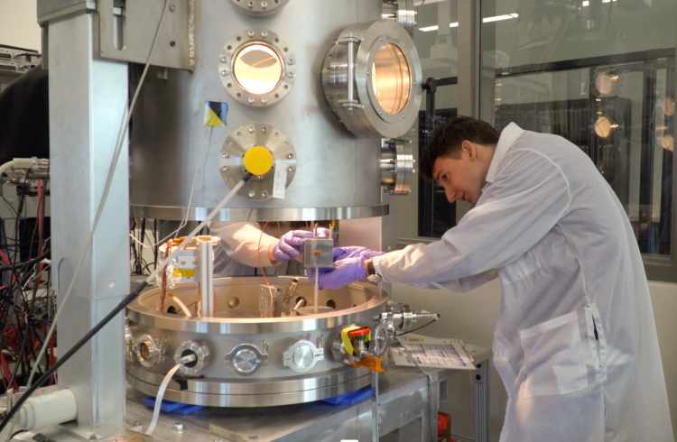 Man wearing a lab coat and gloves makes adjustments inside a large metal chamber