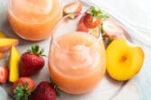 Two glasses of strawberry peach frose on a plate surrounded by slices of strawberries and peaches.