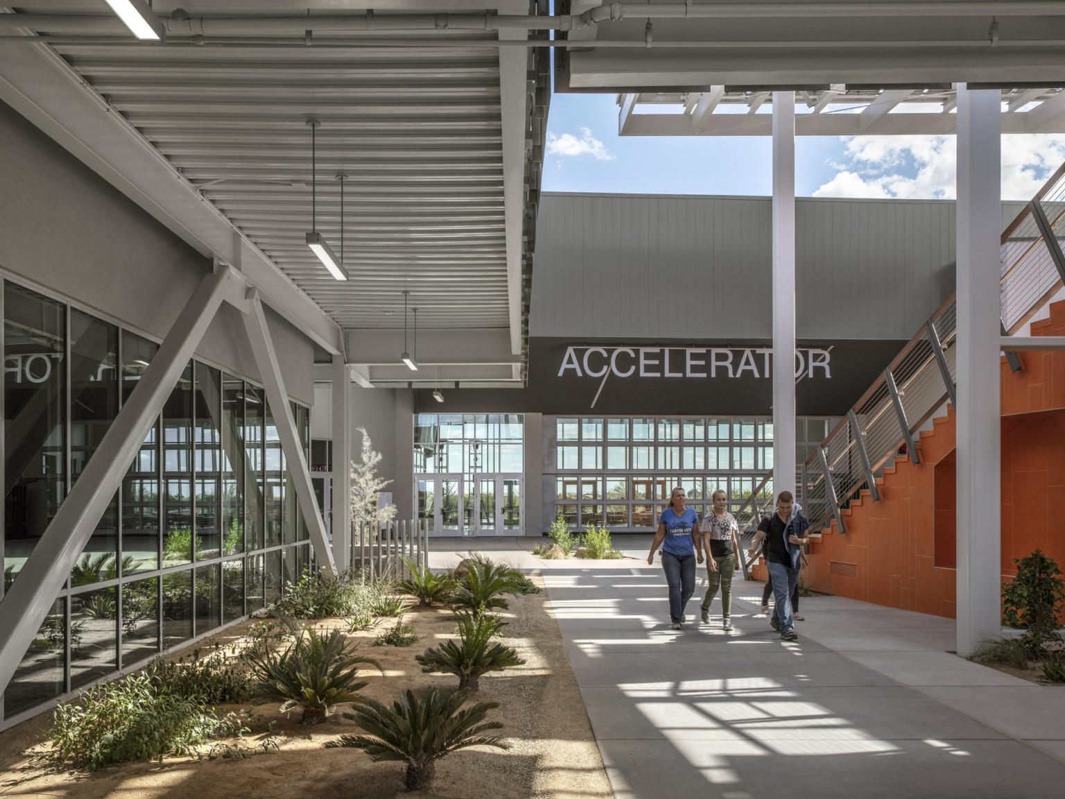 Breezeway to the left of the orange stairs. Behind is a building with a dark roof and parallel walls of double height windows