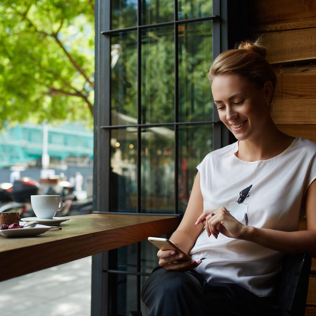 White blonde woman sitting by a window in a cafe smiling while she uses her smartphone.