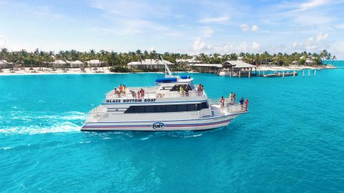 Aerial view of the ocean and a Fury glass bottom boat carrying guests on both top and bottom decks. Behind them is an island with homes and palm trees.