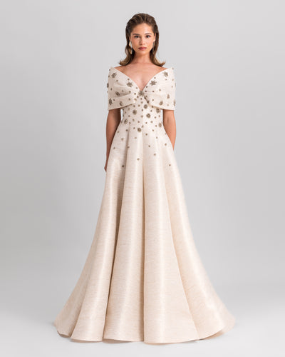 An off-the-shoulders cream evening dress with a bow design, beadings, and a draped skirt