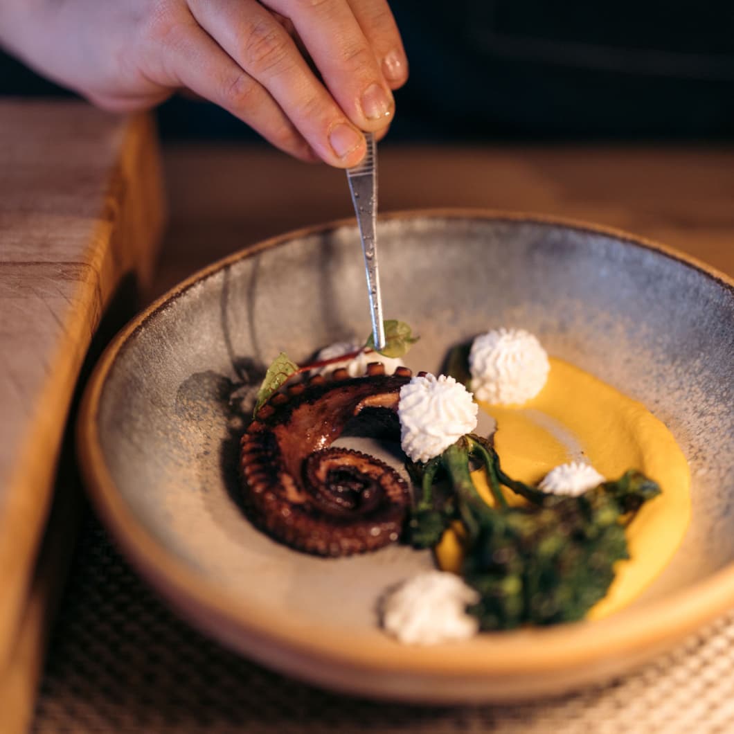 A gourmet plate featuring an artistically arranged seafood over a swipe of yellow sauce, garnished with green leaves and small, white dollops of cream, with a person's hand holding tweezers touching the food.