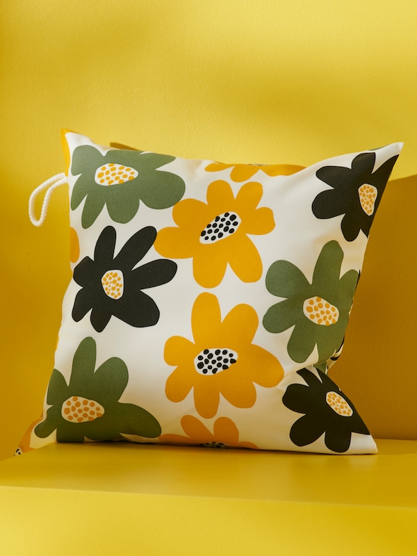 A GULLBERGSÖ cushion cover for outdoor/indoor with a multicolour floral pattern is resting against a bright yellow wall.