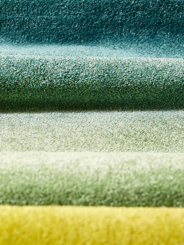 A high pile HOTELLRUM rug with a blue/green and yellow gradient is folded to create a wave effect.