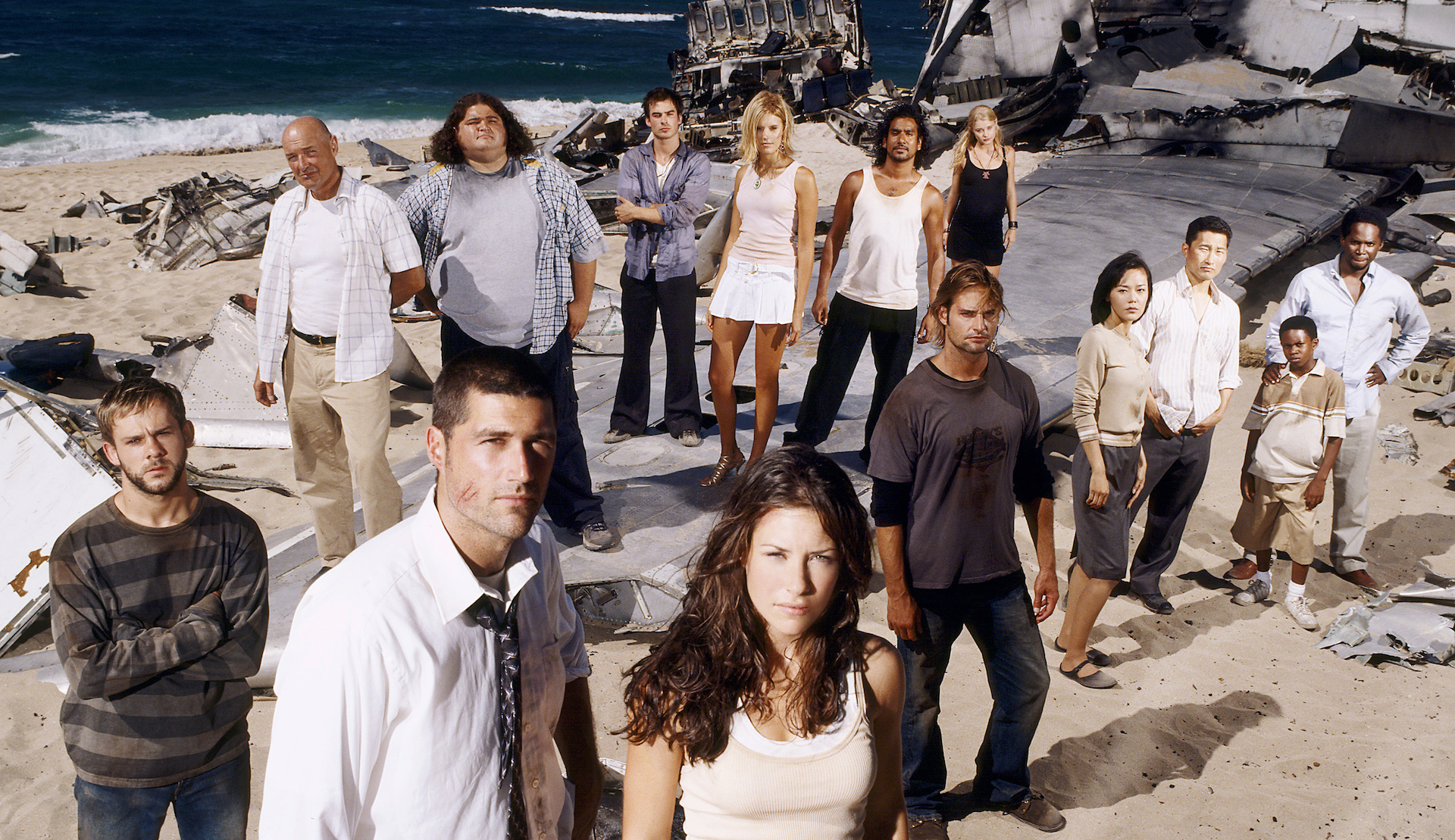 A group of people standing amid plane crash wreckage on a beach and looking directly into camera; promo image for 'Lost'