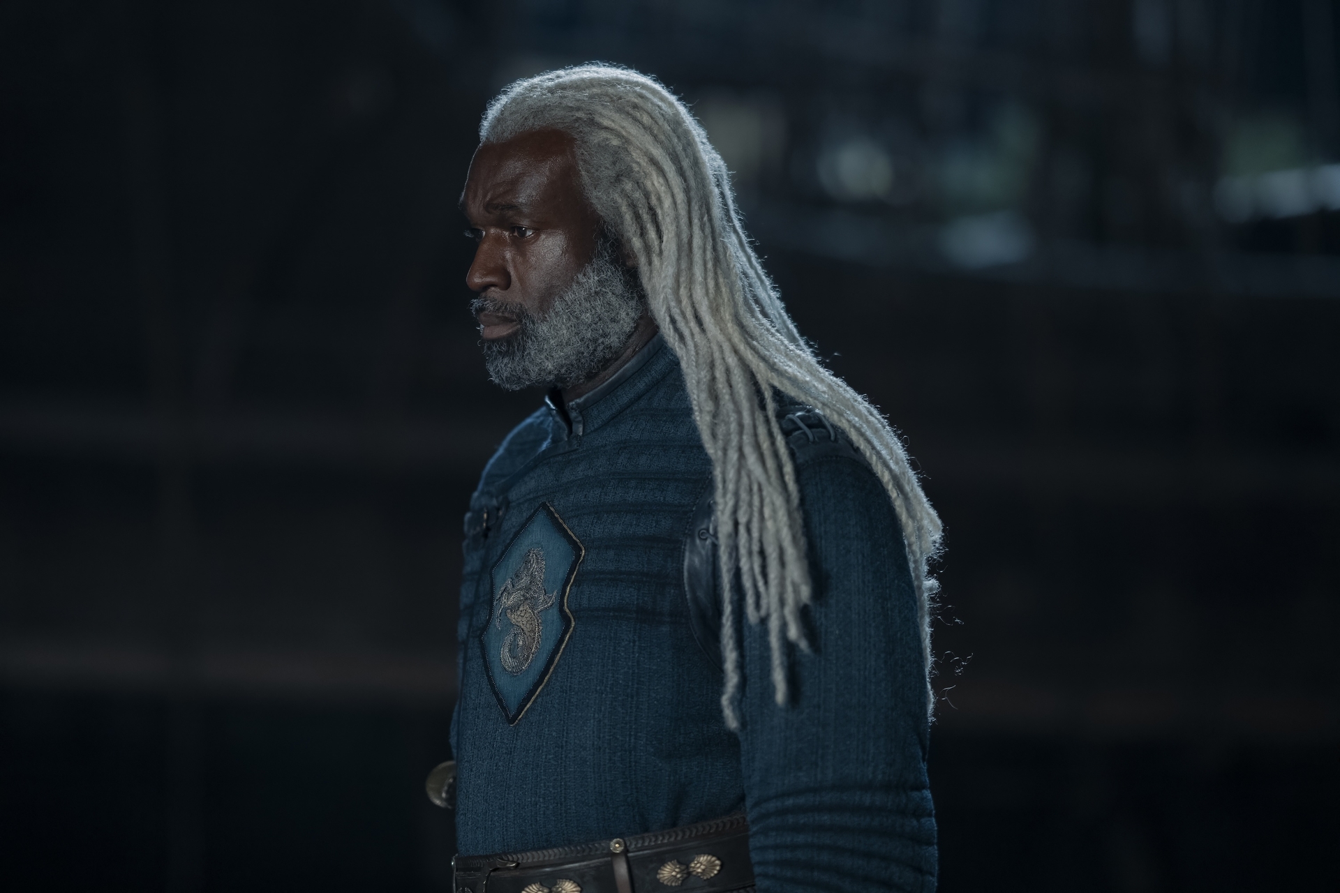 A man in medieval blue clothes with a seahorse sigil on the front and white dreadlocks; Steve Toussaint in 'House of the Dragon'
