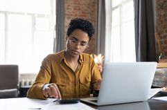 Woman sitting at a desk working on a laptop and entering numbers on a calculator