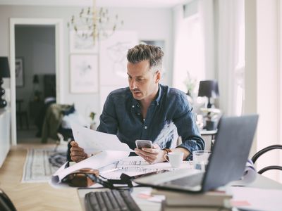 Man in his 40s at his dining room table looking at financial documents and a calculator, with an open laptop nearby