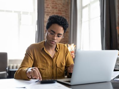 Woman sitting at a desk working on a laptop and entering numbers on a calculator