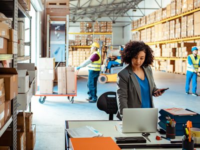 Business manager using laptop in a warehouse with workers moving boxes