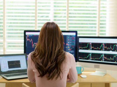 Back of a woman's head as she sits at a computer with 2 monitors and a laptop showing stock charts